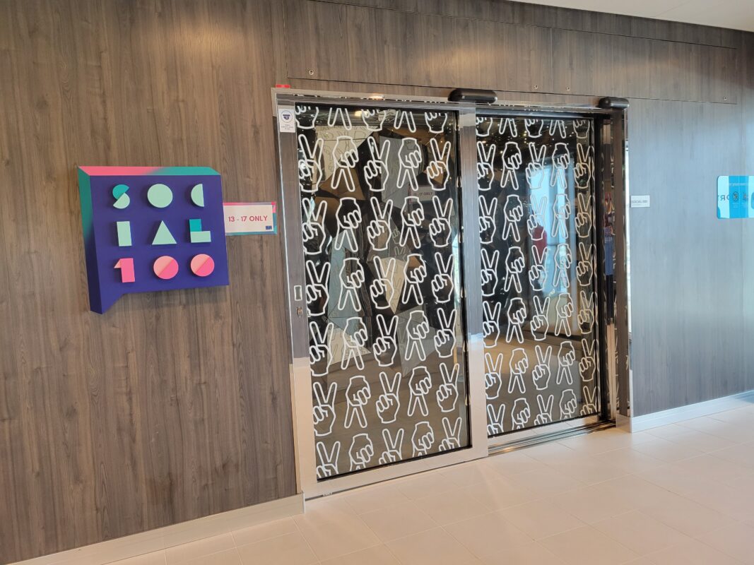 Entrance cruise ship teen club, showing a colorful geometric logo on a wood-paneled wall next to a small "13–17 Only" sign, with glass sliding doors covered in a repeating pattern of white outline hand gesture graphics.