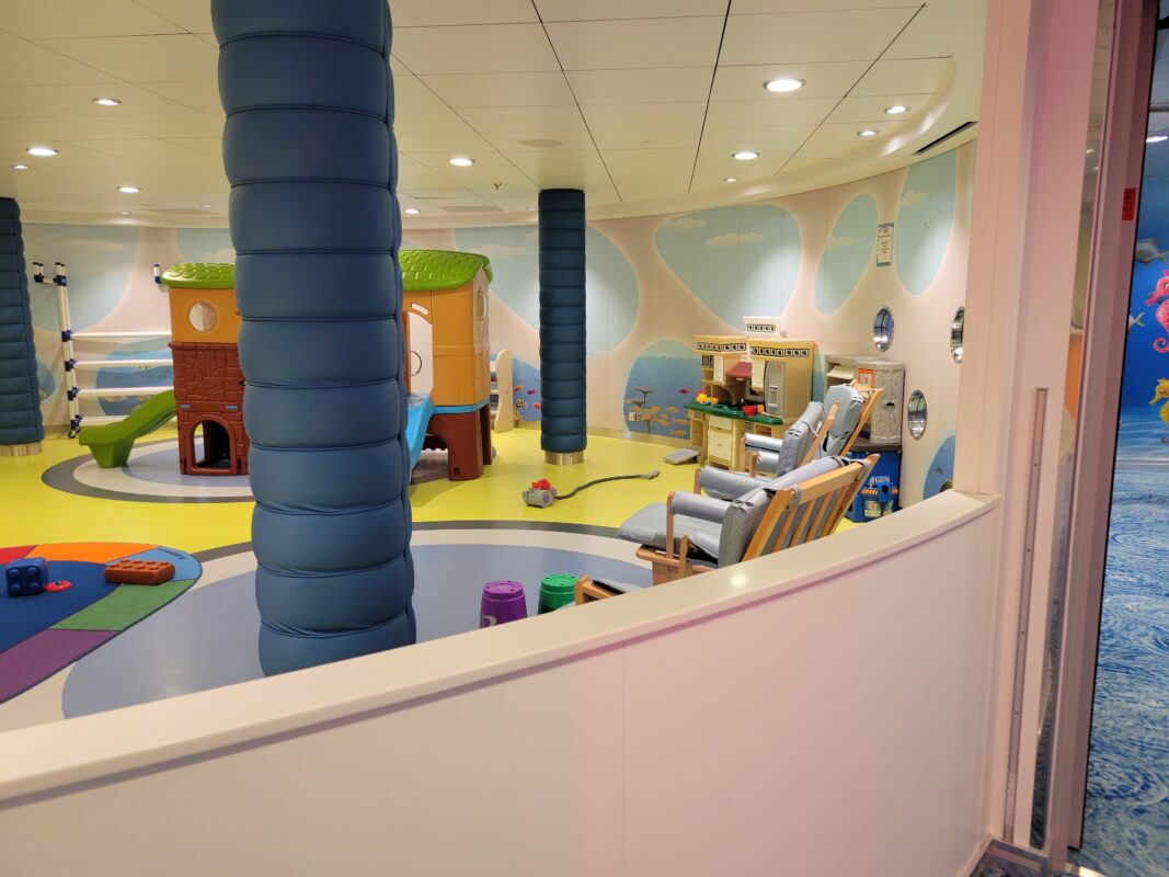 Bright, open cruise ship play space designed for toddlers and parents, featuring a plastic climbing structure with slide, toy kitchen, large building blocks, soft flooring, and whimsical underwater wall murals, with adult seating visible near the play equipment.