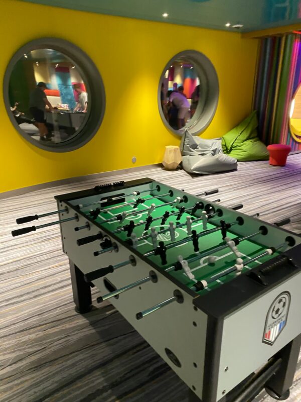 Cruise ship kids' club game room with a foosball table in the foreground, gray and green bean bag chairs against a bright yellow wall, and two large circular porthole-style windows looking into an adjacent room where children are visible playing at a table.