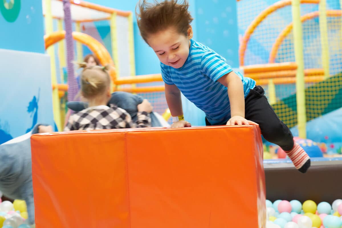 Young boy in a blue striped shirt launching himself over a large orange foam block in a colorful indoor play area, grinning with his hair flying, while other children and a supervising adult are visible in the background amid play equipment and a ball pit.