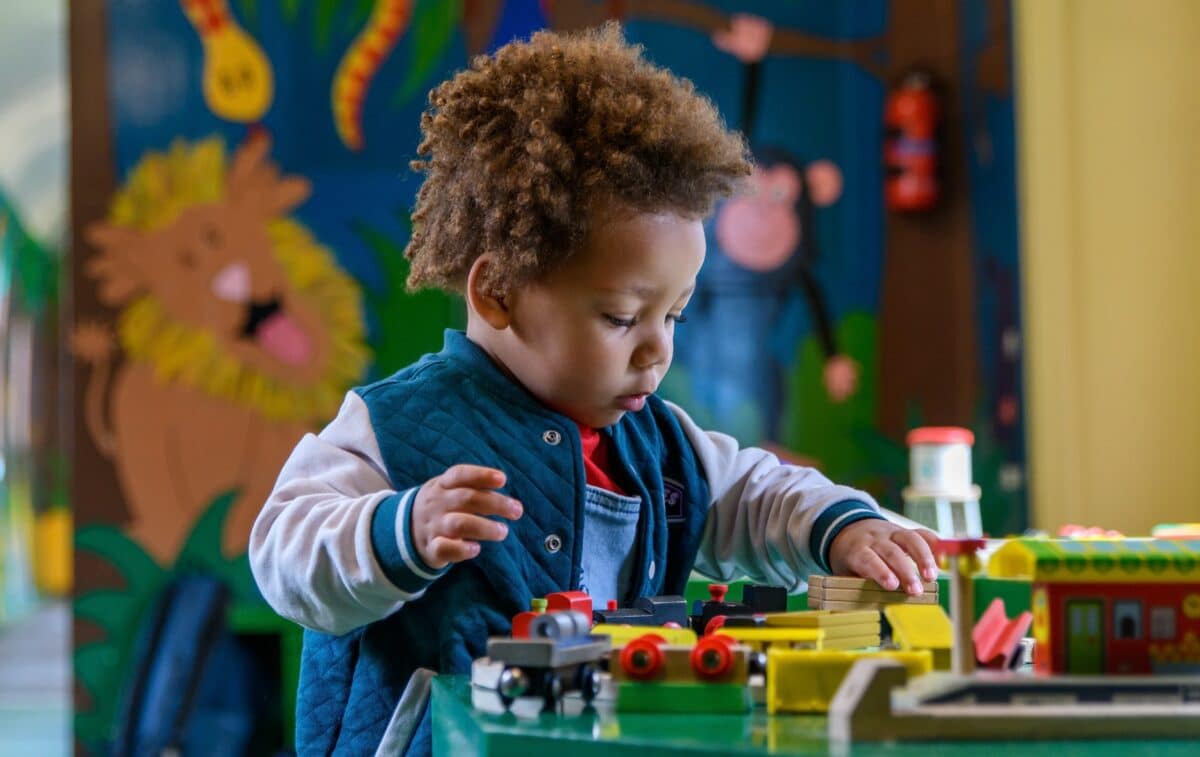 Young bi-racial toddler with curly hair concentrating intently while playing with colorful wooden toy trains and building pieces on a table in a brightly decorated children's play area with a jungle-themed mural visible in the background.