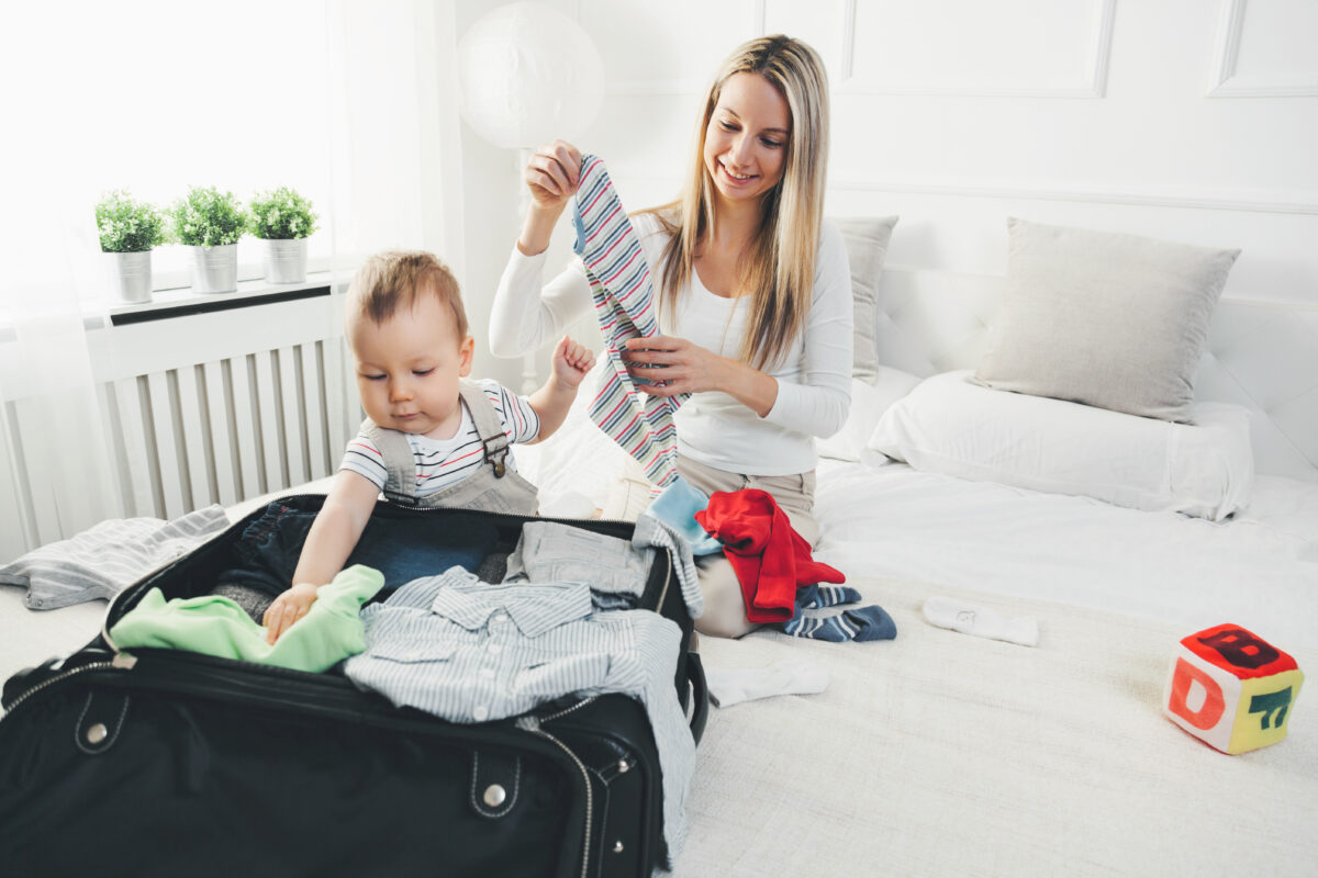 A woman packs baby clothes into a suitcase while a toddler plays among folded shirts and socks.